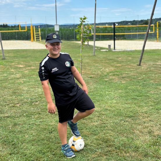 Ein Junge in Sportkleidung lächelt und posiert mit einem Fußball auf einem Außengelände, mit einem Fußballfeld im Hintergrund.