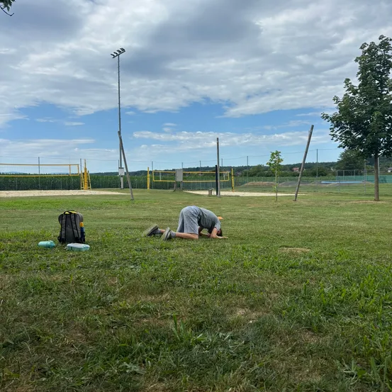 Eine Person ruht sich auf dem Rasen in einem Außenbereich aus. Es gibt einen Rucksack und eine Flasche, die auf dem Gras platziert sind. Ein Pfahl steht in der Ferne. Es gibt zwei Volleyballnetze, die auf dem Boden platziert sind.