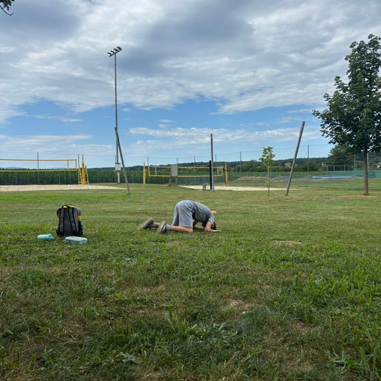 Eine Person ruht sich auf dem Rasen in einem Außenbereich aus. Es gibt einen Rucksack und eine Flasche, die auf dem Gras platziert sind. Ein Pfahl steht in der Ferne. Es gibt zwei Volleyballnetze, die auf dem Boden platziert sind.