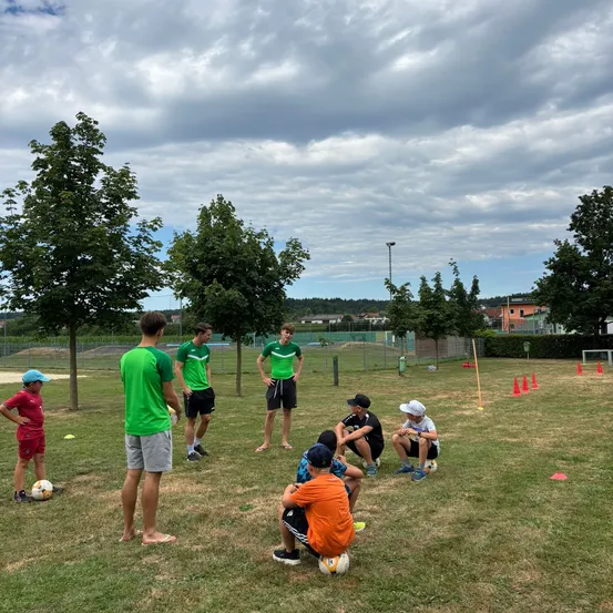 Eine Gruppe von Menschen, darunter junge Jungen, trainiert Fußballübungen auf einem Rasenfeld. Einige stehen und einige sitzen, mit aufgestellten Hütchen und einem Tor. Bäume und ein bewölkter Himmel sind im Hintergrund.