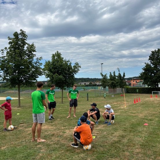 Eine Gruppe von Menschen, darunter junge Jungen, trainiert Fußballübungen auf einem Rasenfeld. Einige stehen und einige sitzen, mit aufgestellten Hütchen und einem Tor. Bäume und ein bewölkter Himmel sind im Hintergrund.