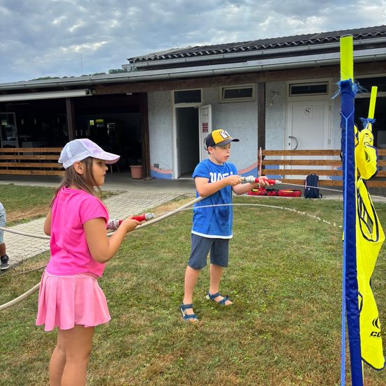 Zwei Kinder spielen mit Wasserpistolen vor einem Gebäude mit bewölktem Himmel. Der Junge trägt eine Baseballkappe, und das Mädchen trägt einen rosa Rock.