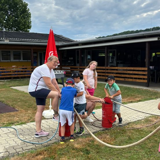 Eine Gruppe von Kindern und eine erwachsene Frau spielen mit einem Feuerlöscher auf dem Gras. Dahinter steht ein Gebäude mit Holzzäunen und einer roten Flagge.