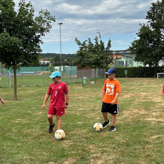 Zwei junge Jungen spielen Fußball in einem Park mit grünen Bäumen und einem bewölkten Himmel. Ein Junge ist in Rot, der andere in Orange.