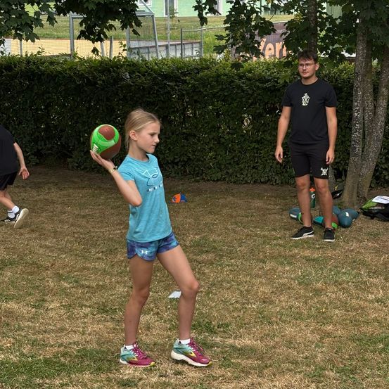Ein Mädchen hält einen Fußball auf einem Rasen. Ein Junge steht hinter ihr und eine Person geht im Hintergrund vorbei.