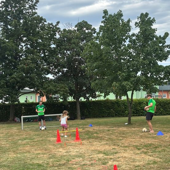 Eine Gruppe von Menschen übt Fußball auf einem offenen Feld mit orangefarbenen Hütchen, einem Tor und einem Fußball. Bäume und Häuser sind im Hintergrund.