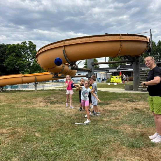 Eine Gruppe von Kindern spielt mit einem Ball auf der Wiese vor einem Wasserrutsche. Eine Frau beobachtet sie.