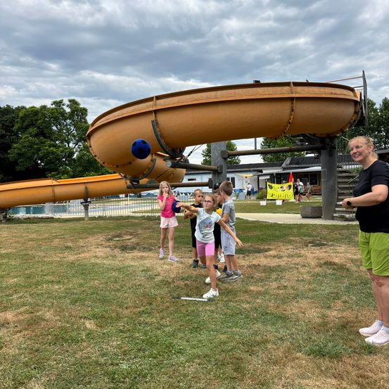 Eine Gruppe von Kindern spielt mit einem Ball auf der Wiese vor einem Wasserrutsche. Eine Frau beobachtet sie.
