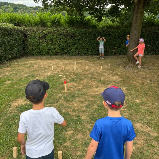 Mehrere Kinder spielen ein Spiel mit Windmühlen auf einer Rasenfläche. Zwei Jungen stehen hinter einem Baum und werfen einen Ball.