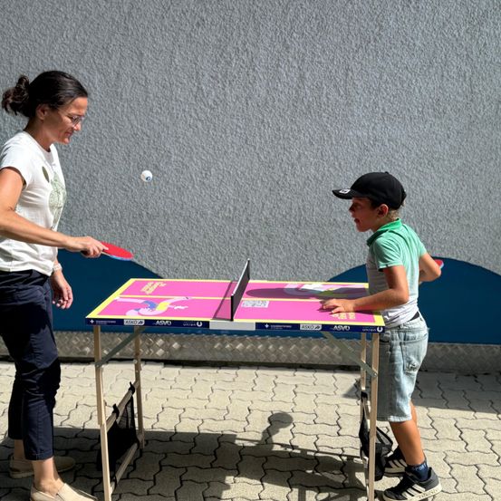 Eine Frau und ein Junge spielen Tischtennis auf einem Tisch im Freien. Der Junge trägt einen Hut und Turnschuhe, während die Frau eine Brille und Turnschuhe trägt. Der Ball ist in der Luft, und der Junge schaut ihn an. Der Tisch hat ein farbenfrohes Design.