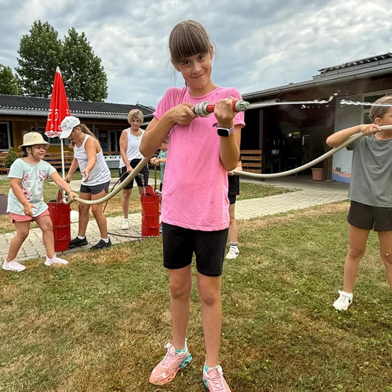Kinder spielen mit einem Wasserschlauch, den sie in den Händen halten. Ein Mädchen mit einem rosa Shirt steht vor zwei roten Eimern.