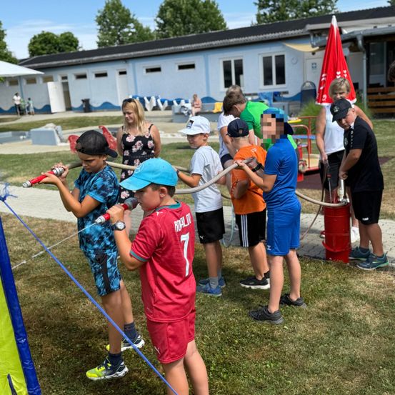 Kinder halten Wasserschläuche und sprühen Wasser nach draußen. Ein Junge trägt einen blauen Hut. Dahinter sind ein Gebäude und ein roter Regenschirm zu sehen.
