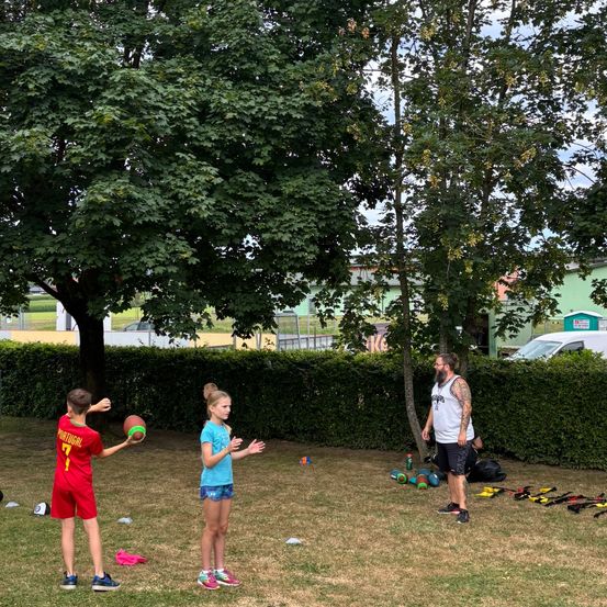Eine Gruppe von Kindern und einem Erwachsenen spielt ein Ballspiel auf einem Grasfeld, umgeben von Bäumen und Büschen. Der Erwachsene steht in der Nähe von Fitnessgeräten.