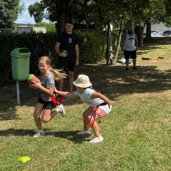 Zwei junge Mädchen spielen draußen ein Fußballspiel. Eine läuft und hält einen Fußball. Ein Mann und eine Frau stehen in der Nähe. Bäume und Büsche sind im Hintergrund.