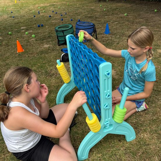 Zwei Mädchen spielen an einem sonnigen Tag ein Riesen-Connect-Four-Spiel. Ein Mädchen konzentriert sich auf das Spiel, während das andere zuschaut.