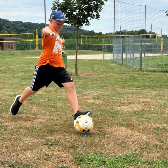 Ein Junge in einem orangefarbenen T-Shirt und schwarzen Shorts tritt einen Fußball auf einem offenen Feld. Hinter ihm befinden sich Metallzäune und Bäume.