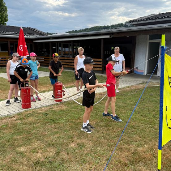 Eine Gruppe von Kindern und Erwachsenen spielt mit Wasserpistolen. Sie sind draußen, wobei zwei Erwachsene sie beaufsichtigen. Im Hintergrund befindet sich ein Gebäude, und die Kinder tragen verschiedenfarbige Shirts und Shorts.