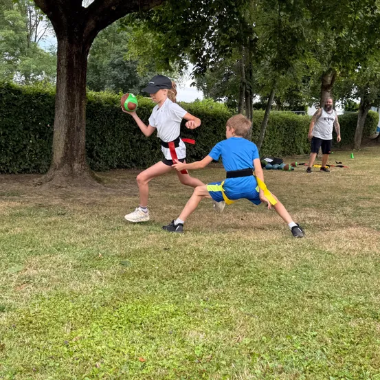 Ein Mädchen hält einen Ball und läuft, während ein Junge versucht, sie in einem Park mit grünen Hecken und Bäumen zu fangen. Ein Mann steht in der Nähe.