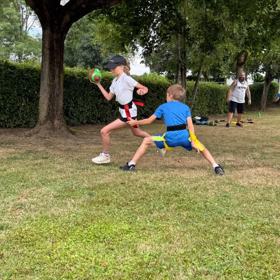 Ein Mädchen hält einen Ball und läuft, während ein Junge versucht, sie in einem Park mit grünen Hecken und Bäumen zu fangen. Ein Mann steht in der Nähe.