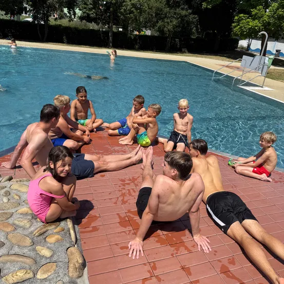 Eine Gruppe von Kindern und Erwachsenen lounget am Rande eines Schwimmbeckens, einige sitzen auf den Kacheln und andere stehen oder knien. Das Becken ist mit klarem blauem Wasser gefüllt, und eine Person schwimmt im Hintergrund.