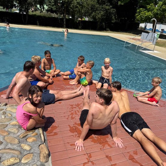 Eine Gruppe von Kindern und Erwachsenen lounget am Rande eines Schwimmbeckens, einige sitzen auf den Kacheln und andere stehen oder knien. Das Becken ist mit klarem blauem Wasser gefüllt, und eine Person schwimmt im Hintergrund.
