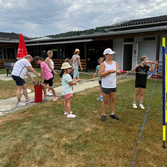 Eine Gruppe von Menschen, einschließlich Kindern, ist auf einem Rasenfeld mit einer Wasseraktivität beschäftigt und verwendet Schläuche, die mit einem roten Wassertank verbunden sind. Sie befinden sich in der Nähe eines Gebäudes mit einem geschlossenen Eingang.