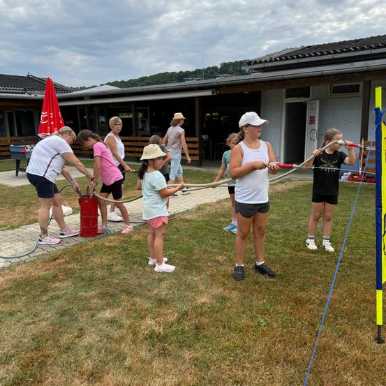 Eine Gruppe von Menschen, einschließlich Kindern, ist auf einem Rasenfeld mit einer Wasseraktivität beschäftigt und verwendet Schläuche, die mit einem roten Wassertank verbunden sind. Sie befinden sich in der Nähe eines Gebäudes mit einem geschlossenen Eingang.