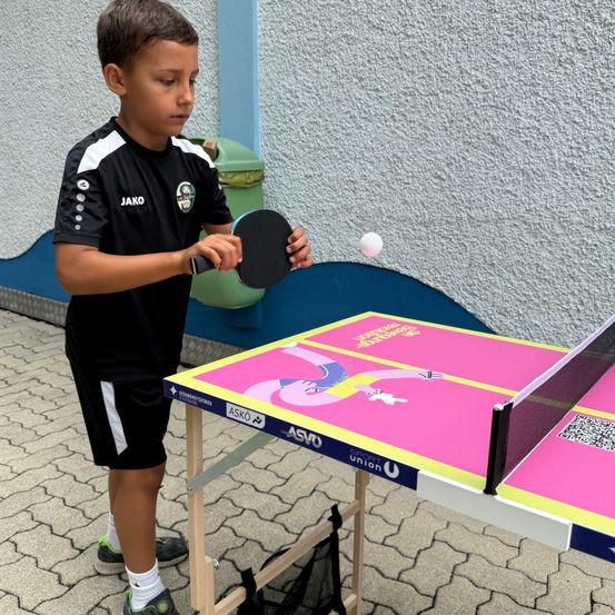 Ein junger Junge in einem schwarzen Sportshirt spielt Tischtennis und schlägt einen Ball mit einem Schläger. Der Tisch hat ein buntes Design.