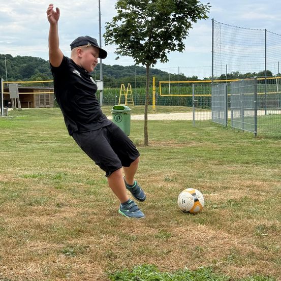 Ein Junge kickt einen Fußball auf einem Rasenfeld neben einem Baum und einem grünen Mülleimer.
