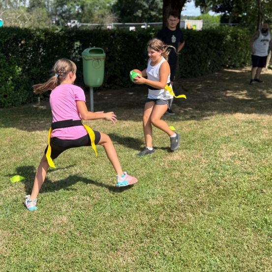Zwei Mädchen spielen auf einem Grasfeld. Eine hält einen grünen Ball, beide tragen gelbe Bänder. Ein Mülleimer und ein Mann im Hintergrund.