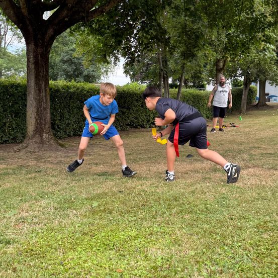 Zwei Jungen spielen auf einem Rasenfeld Fußball, während ein Trainer zusieht. Ein Junge hält einen Football, während der andere versucht, ihn zu blockieren. Bäume und eine Hecke umgeben das Gebiet.