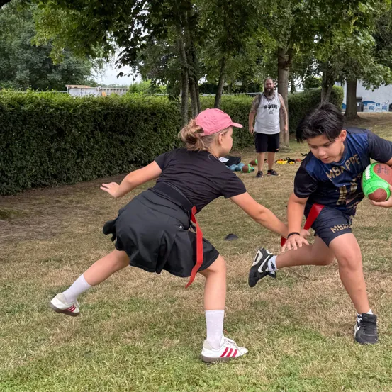 Ein junges Mädchen in einem schwarzen Shirt und Rock tacklet einen Jungen in einem blauen Trikot, der einen Rugby-Ball trägt. Dahinter steht ein Mann mit Bart in der Nähe einiger Bäume.