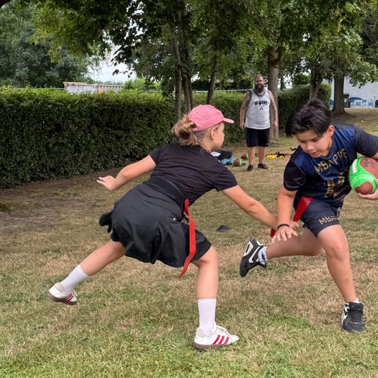 Ein junges Mädchen in einem schwarzen Shirt und Rock tacklet einen Jungen in einem blauen Trikot, der einen Rugby-Ball trägt. Dahinter steht ein Mann mit Bart in der Nähe einiger Bäume.