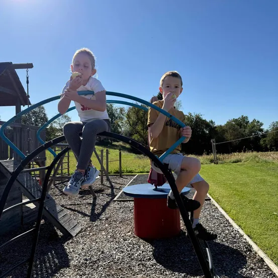 Zwei Kinder sitzen auf einer geschwungenen Metallstruktur in einem Spielplatz und essen Äpfel. Die Struktur ist blau und schwarz, und der Hintergrund ist ein Grasfeld mit Bäumen.