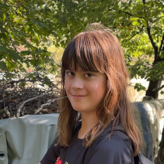 Ein junges Mädchen mit braunen Haaren und Pony lächelt in einem Waldgebiet für die Kamera.