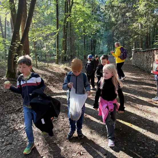 Eine Gruppe von Kindern geht auf einem Waldweg, einige tragen Taschen und Jacken. Ein Kind hält einen Apfel, während andere nach unten oder vor sich schauen.