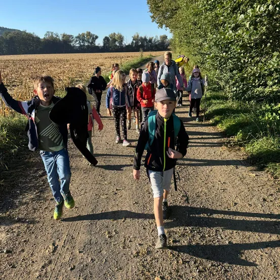 Eine Gruppe von Kindern und Erwachsenen geht auf einem Feldweg, einige tragen Rucksäcke und Turnschuhe.