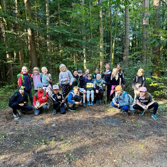 Eine Gruppe von Kindern und Erwachsenen posiert für ein Foto im Wald, einige sitzen auf dem Boden und andere stehen hinter ihnen, alle tragen Rucksäcke und Mützen.