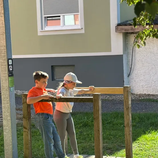 Zwei Kinder, ein Junge in Orange und ein Mädchen in Weiß, stehen auf einer Holzstruktur in einem Grasbereich vor einem Haus.