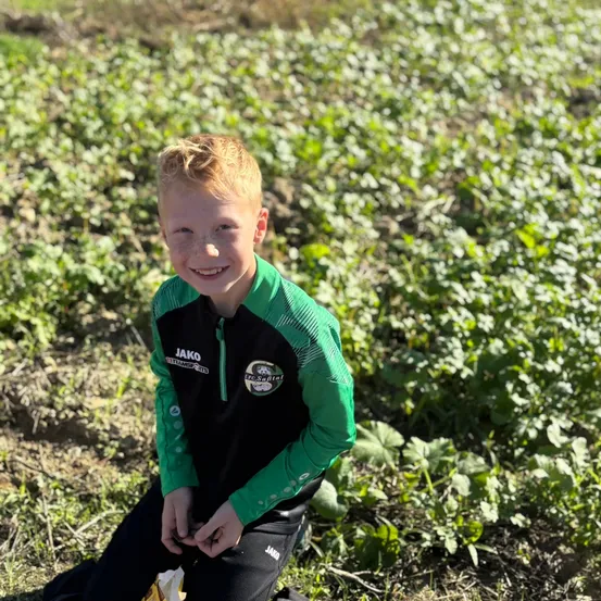 Ein junger Junge mit blonden Haaren kniet in einem Feld grüner Pflanzen und trägt eine grün-schwarze Sportjacke mit einem Logo auf der Brust.