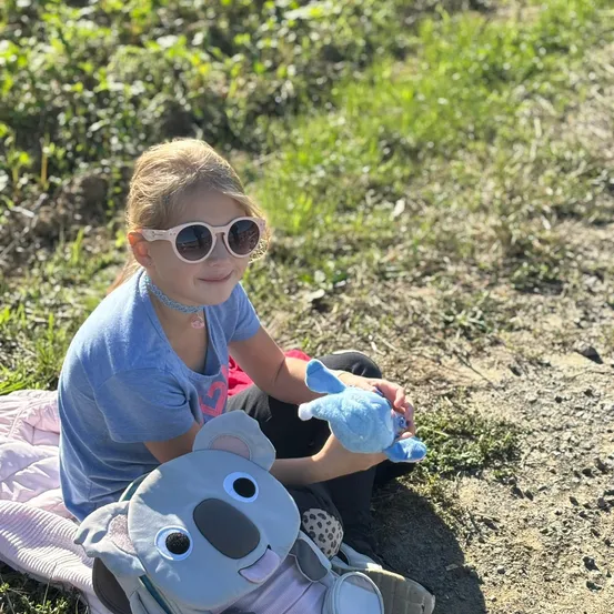 Ein junges Mädchen mit blonden Haaren und Sonnenbrille sitzt auf dem Boden und hält ein blaues Koala-Plüschtier.