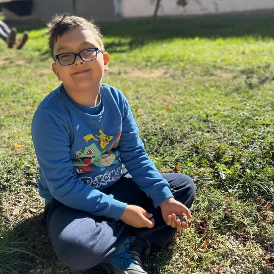 Ein junger Junge mit Brille sitzt auf dem Rasen, lächelt und hält einen kleinen Stein. Er trägt ein blaues Sweatshirt mit einem Pokemon-Charakter.