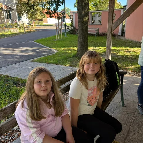 Zwei junge Mädchen sitzen auf einer Holzbank im Freien, eines trägt eine rosa Jacke und das andere ein weißes T-Shirt mit einem Cartoon-Charakter. Sie lächeln und scheinen für ein Foto zu posieren.