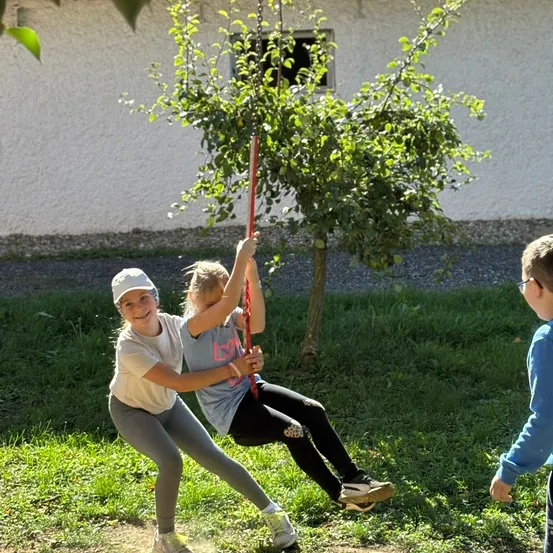 Zwei Mädchen spielen auf einer Schaukel im Hof, wobei ein Mädchen die Schaukel hält und das andere Mädchen darauf sitzt. Ein Junge steht in der Nähe.