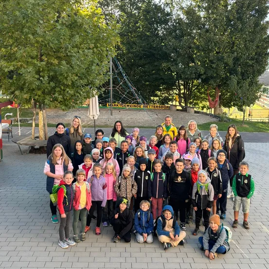 Eine Gruppe von Kindern und Erwachsenen posiert für ein Foto in einem Park mit Bäumen und einem Spielplatz im Hintergrund.