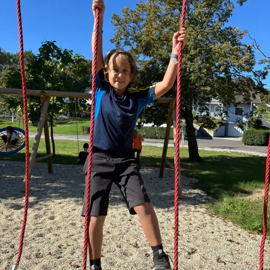 Ein kleiner Junge klettert an einem roten Seilschaukel in einem Spielplatz, mit einem Haus und Bäumen im Hintergrund.