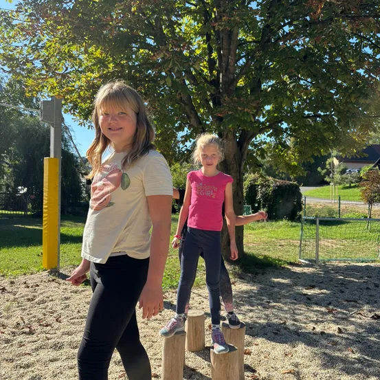 Zwei junge Mädchen stehen auf Holzstämmen in einem Park, lächeln und posieren für ein Foto. Hinter ihnen steht ein Baum und ein Zaun.