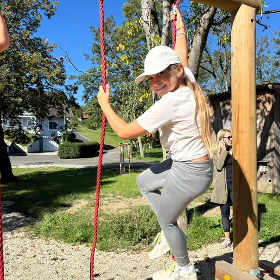 Ein junges Mädchen klettert an einem roten Seil in einem Park. Sie trägt eine weiße Mütze und graue Leggings. Eine andere Frau beobachtet in der Nähe.