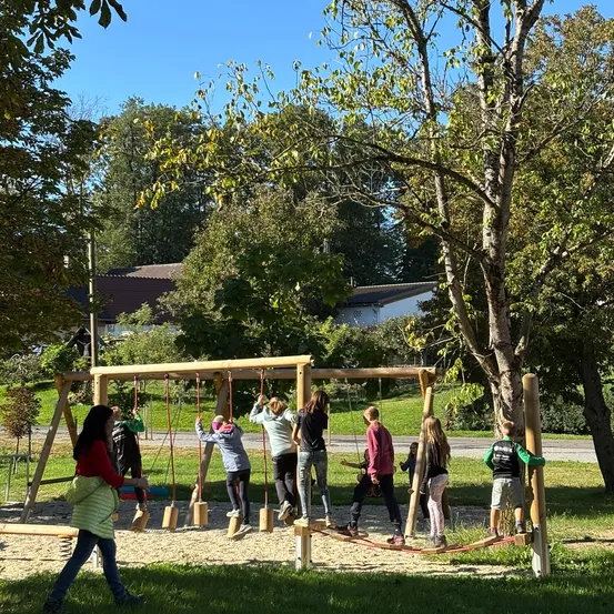 Eine Gruppe von Kindern spielt auf einer hölzernen Spielplatzstruktur mit Schaukeln und einer Balancestange unter einem klaren Himmel.