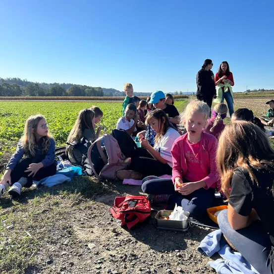 Eine Gruppe von Kindern und Erwachsenen sitzt auf dem Boden in einem Feld, einige essen und andere beobachten. Ein klarer blauer Himmel und entfernte Bäume sind sichtbar.
